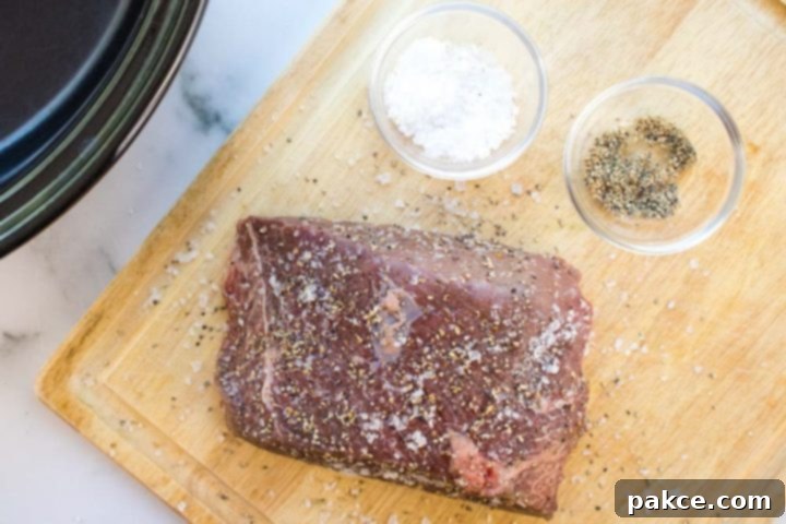 Crockpot Roast Beef & Gravy An uncooked beef roast sprinkled with salt and pepper on a wooden cutting board next to a slow cooker. There is a small glass bowl of salt and a small glass bowl of pepper on the cutting board next to the roast.