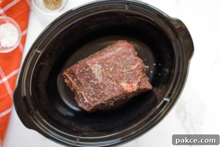 Crockpot Roast Beef & Gravy Uncooked beef roast in a slow cooker on a white marble counter. On the left side of the photo is an orange napkin with white stripes. On the napkin is a small glass bowl with salt. Next to the napkin is small glass bowl with pepper.