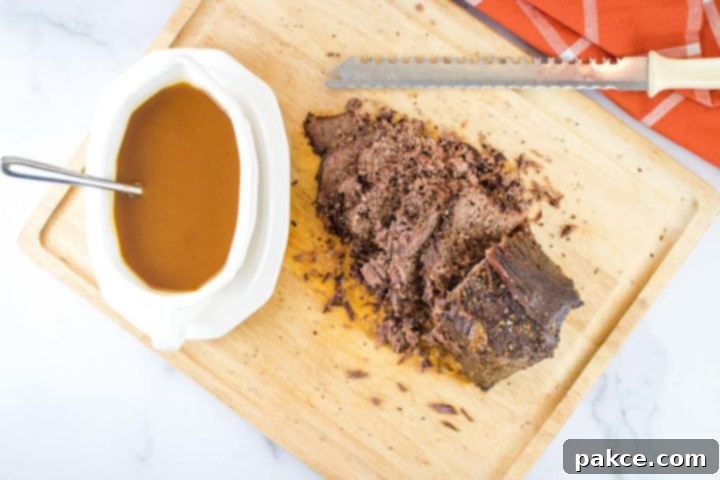Crockpot Roast Beef & Gravy White gravy bowl on a white platter. The bowl has brown gravy and a spoon. The platter is sitting on a wooden cutting board next to a beef roast that has been half-sliced with the serrated knife at the top of the photo. There is an orange napkin with white stripes in the top right corner of the photo.