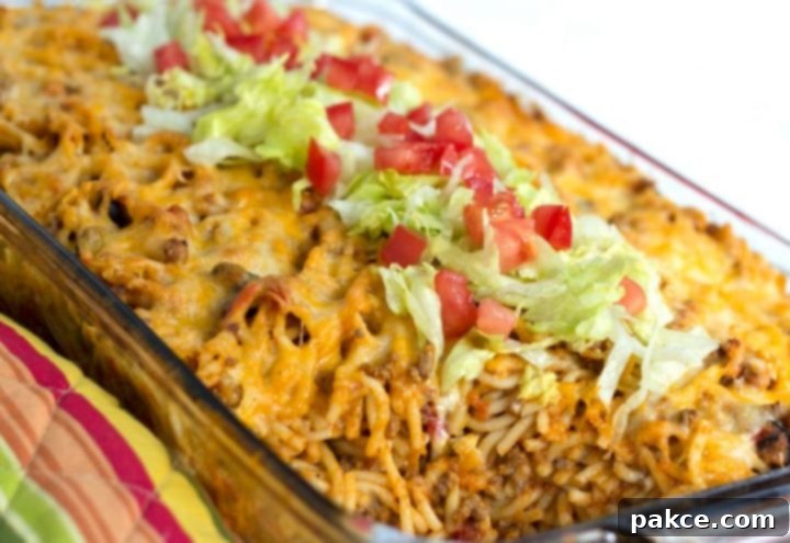Clear casserole dish with spaghetti noodles, ground beef, and melted cheese, topped with shredded lettuce and diced tomatoes.