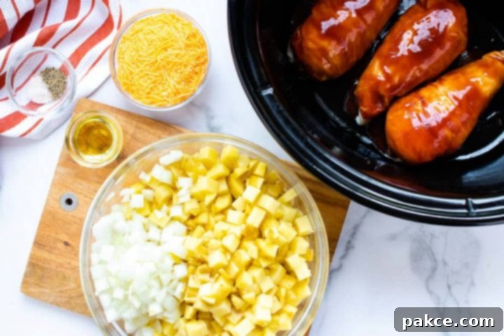 Small bowl with salt and pepper sitting on an orange and white striped napkin, a bowl of shredded cheddar cheese, a slow cooker with three chicken breasts in barbecue sauce, a wooden cutting board a bowl of diced potatoes and onion, and a small bowl with oil.