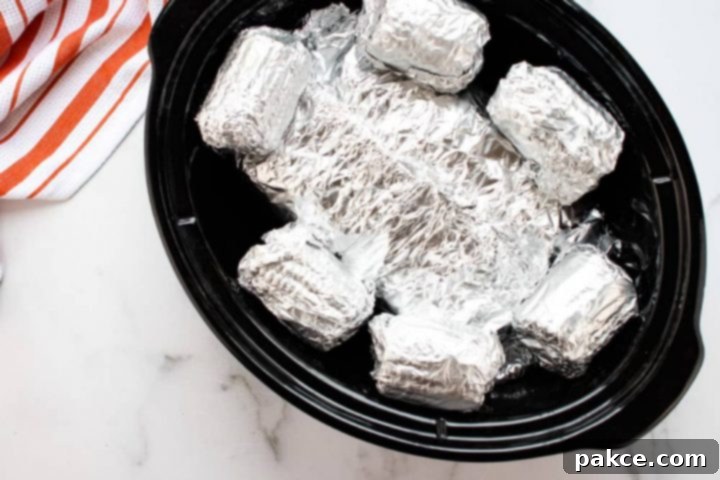 Slow cooker with six small corn cobs wrapped in foil and another foil packet beneath them.
