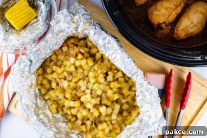 A bowl with foil and a small corn cob, open foil packet with diced potatoes, a slow cooker with chicken breasts. The potatoes are on a wooden cutting board with a basting brush and a set of tongs.