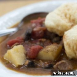Hearty Beef Stew with Fluffy Dumplings 2 White bowl of beef stew with two dumplings on top. Stew consists of brown broth, diced beef, chopped potatoes, and stewed tomatoes. There is a spoon in the bowl on the left side.