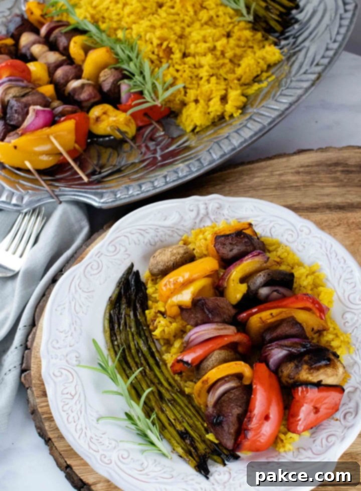 Grilled beef and vegetable kabobs with vibrant red, yellow, and orange bell peppers, mushrooms, and red onions, served over golden yellow rice with grilled asparagus on a white plate, alongside a platter of additional kabobs.