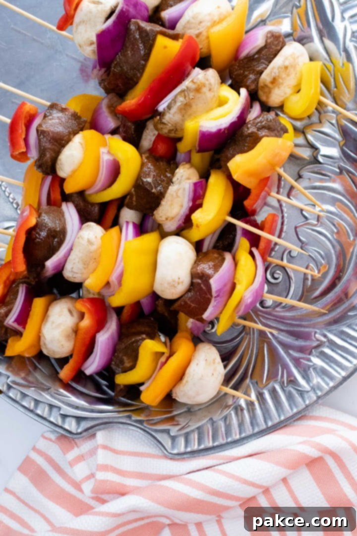 Uncooked beef and vegetable kabobs featuring marinated beef chunks alternated with vibrant red, yellow, and orange bell peppers, whole mushrooms, and red onion pieces, neatly arranged on a pewter platter, ready for grilling.