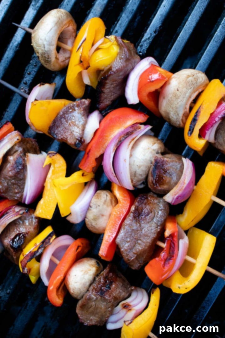 Vibrant beef and vegetable kabobs, featuring red, yellow, and orange bell peppers, mushrooms, and red onions alternating with marinated beef, cooking on a hot grill, showing grill marks and sizzling. 