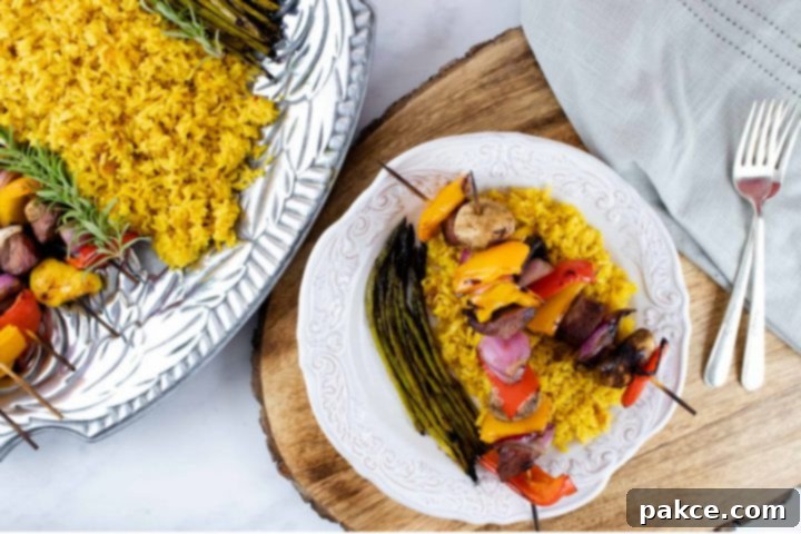 A beautifully plated meal featuring grilled beef and vegetable kabobs arranged over a bed of yellow rice, accompanied by grilled asparagus, all served on a pristine white plate with a silver fork. A larger serving platter with more kabobs, rice, and asparagus is visible in the background.