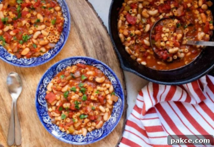 The left of the photo is two blue and white soup bowls, each filled with Pasta e Fagioli, a soup made with pasta, tomatoes, chopped celery, onion, and parsley, cannellini beans, and tomato sauce. There are two spoons next to the bowls. On the right side of the bowls is a cast iron skillet with Pasta e Fagioli. There's an orange and white striped towel on the counter to the right of the skillet, and a ladle scooping soup out of the skillet.
