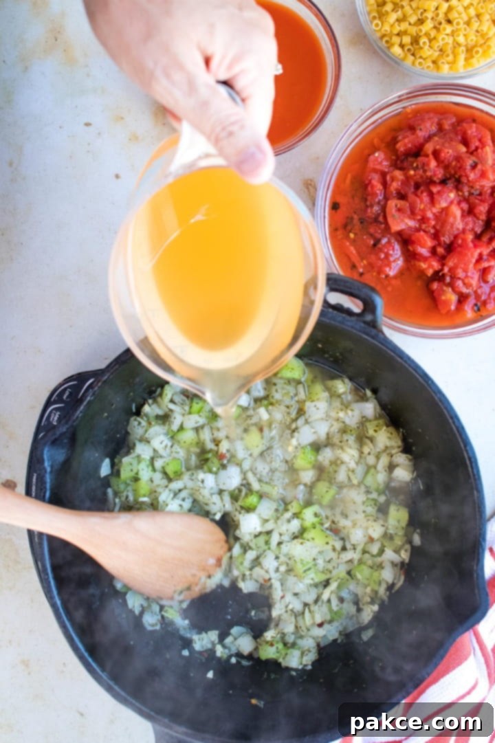 Pasta e Fagioli A measuring cup of broth is being poured into a cast iron stew pot of chopped celery and onions. A wooden spoon is stirring the contents of the pan. At the top right of the photo is a clear glass bowl with roasted tomatoes in tomato juice.