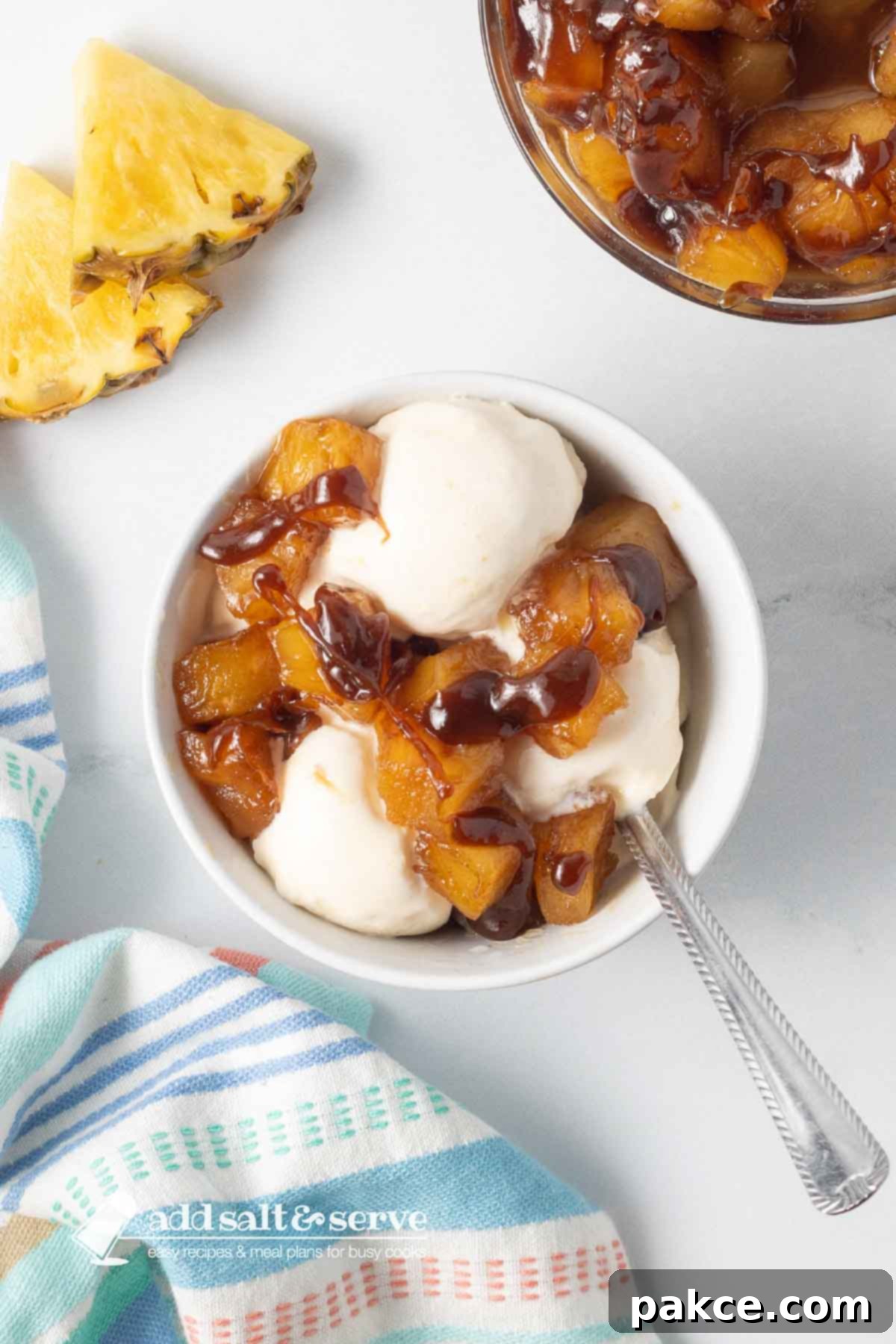 Effortless 3-Ingredient Caramelized Pineapple 5 Bowl of caramelized pineapple chunks and vanilla ice cream drizzled with caramel sauce beside a tea towel and sliced pineapple on a counter.