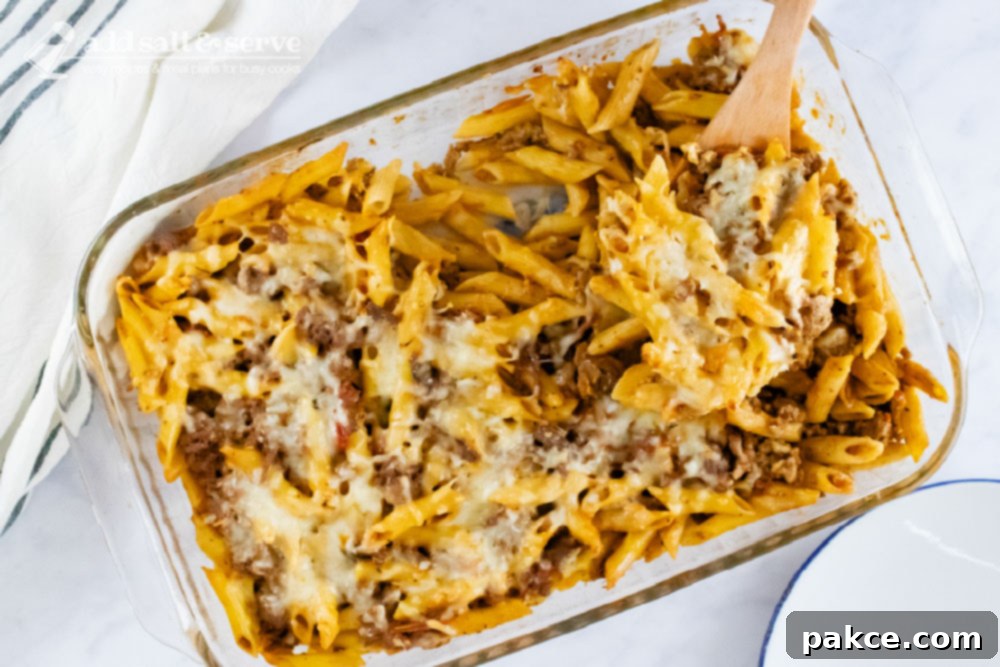 Overhead view of penne pasta bake topped with melted cheese in a glass pan with a spoonful scooped out and held toward the camera.