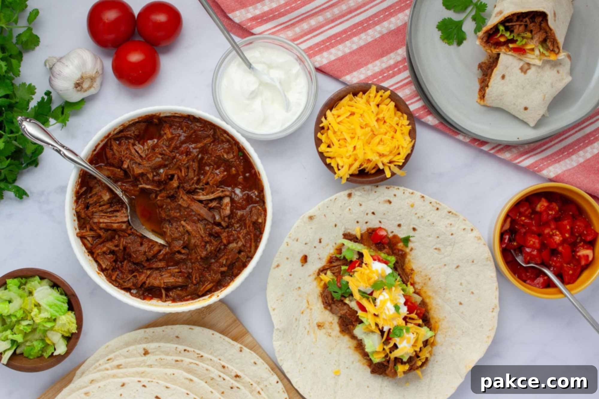 Overhead view of slow cooker beef burrito meat in a bowl surrounded by garnishes, tortillas, and a plate with a half-assembled burrito. A spread of fresh ingredients for a DIY burrito bar.