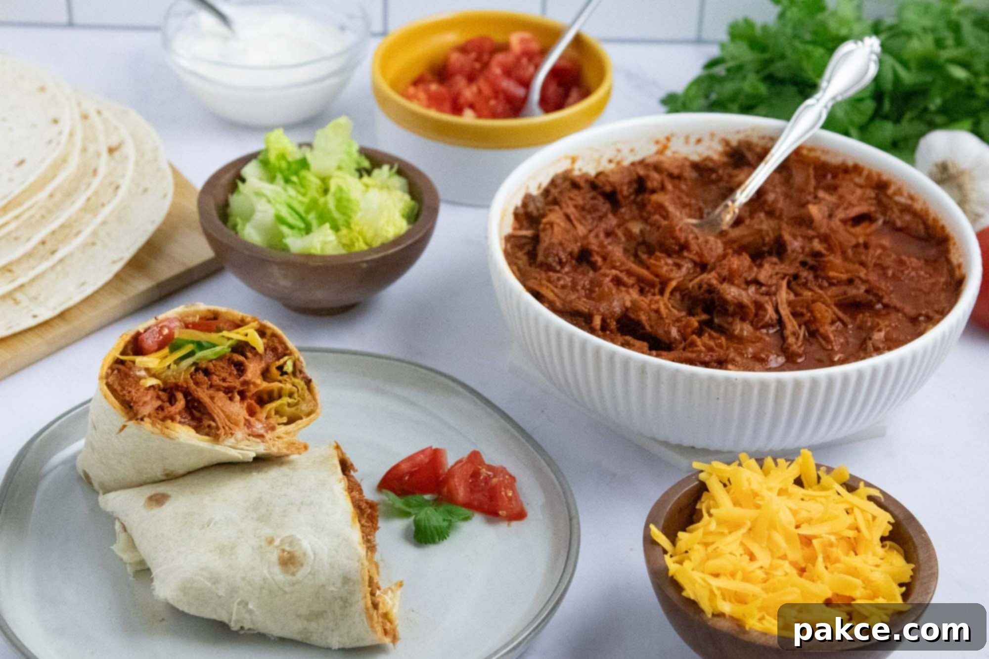Slow Cooker Shredded Beef Burritos 7 Overhead view of slow cooker beef burrito meat in a bowl surrounded by garnishes, tortillas, and a plate with an assembled burrito.