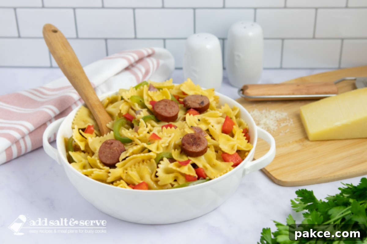 Bowtie pasta with red and green bell peppers, red onion, and sliced kielbasa with a spoon in a serving dish beside fresh parsley and Parmesan cheese.