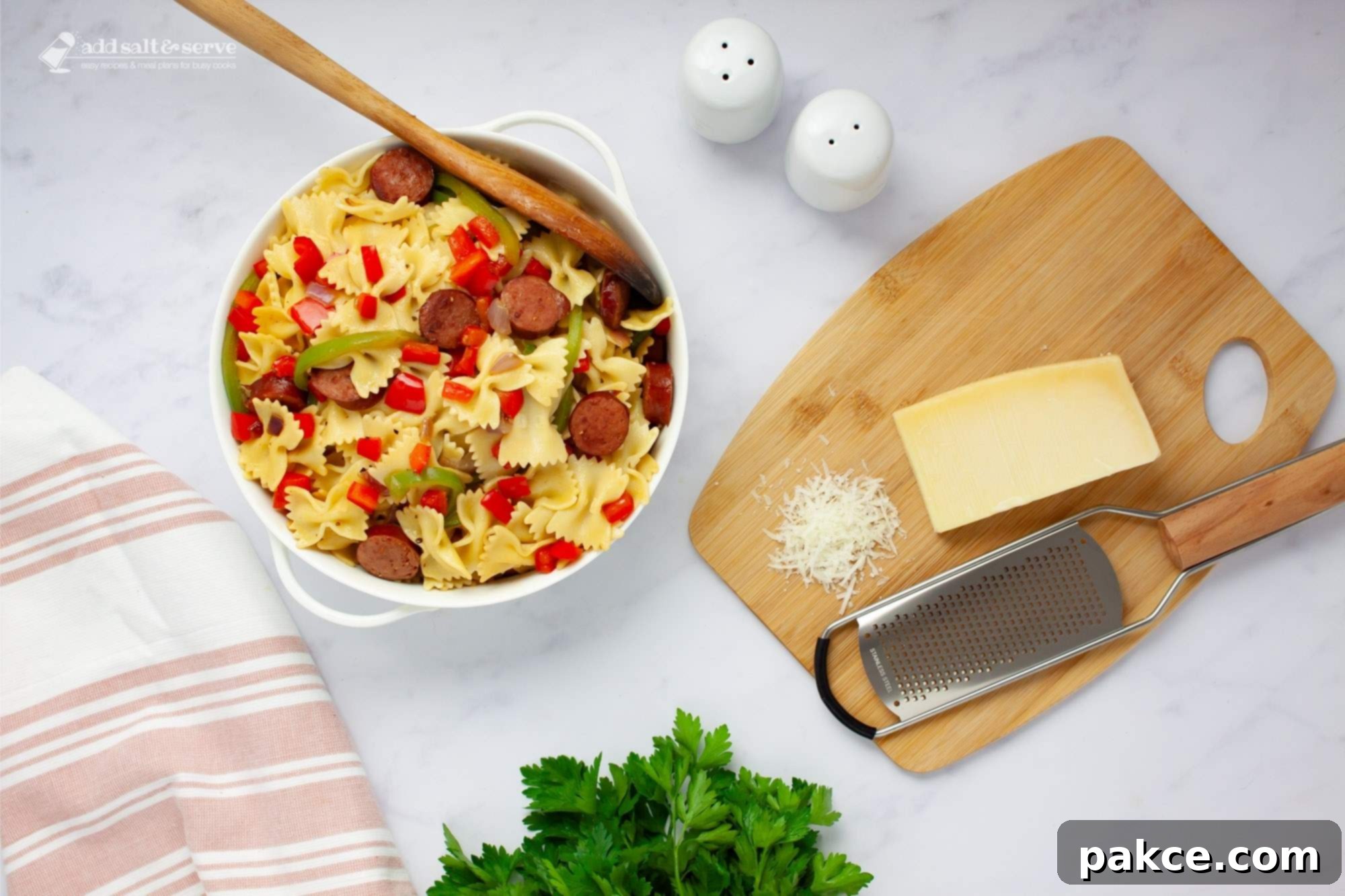 Hearty 20 Minute Kielbasa Pepper Pasta 6 Bowtie pasta with red and green bell peppers, red onion, and sliced kielbasa with a spoon in a serving dish beside fresh parsley and Parmesan cheese.