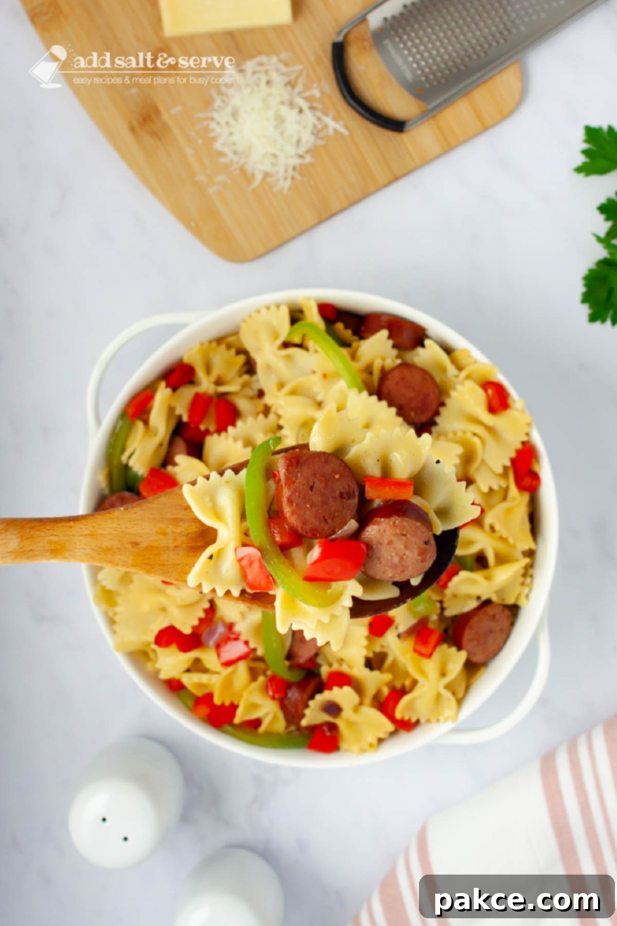 Hearty 20 Minute Kielbasa Pepper Pasta 7 Bowtie pasta with red and green bell peppers, red onion, and sliced kielbasa with a spoon in a serving dish beside fresh parsley and Parmesan cheese.