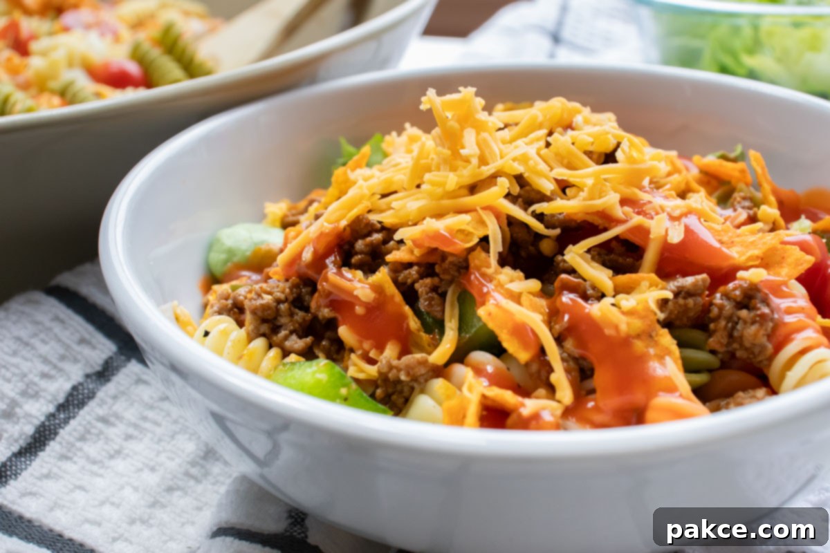 Bowl of seasoned ground beef, spiral pasta, lettuce, halved tomatoes, nacho cheese Doritos, and French salad dressing with shredded cheddar cheese on top.