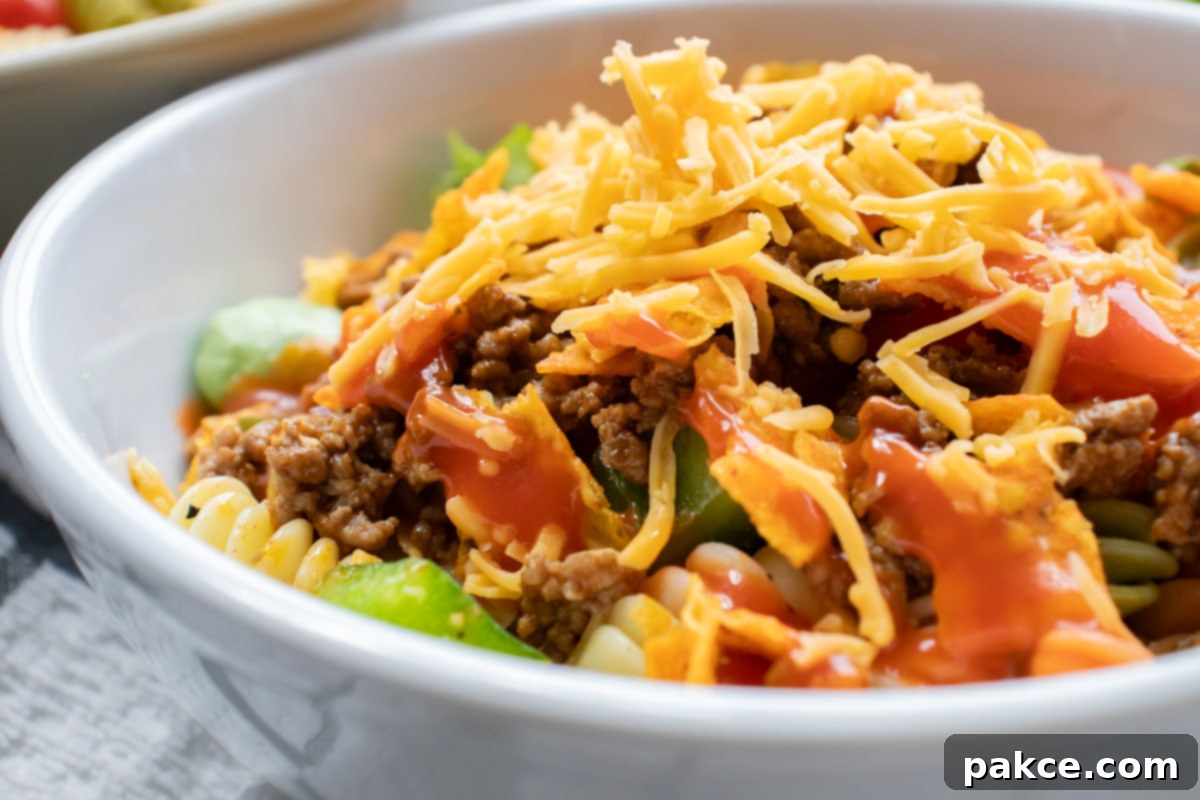 Taco Mac Salad Bowl of seasoned ground beef, spiral pasta, lettuce, halved tomatoes, nacho cheese Doritos, and French salad dressing with shredded cheddar cheese on top.