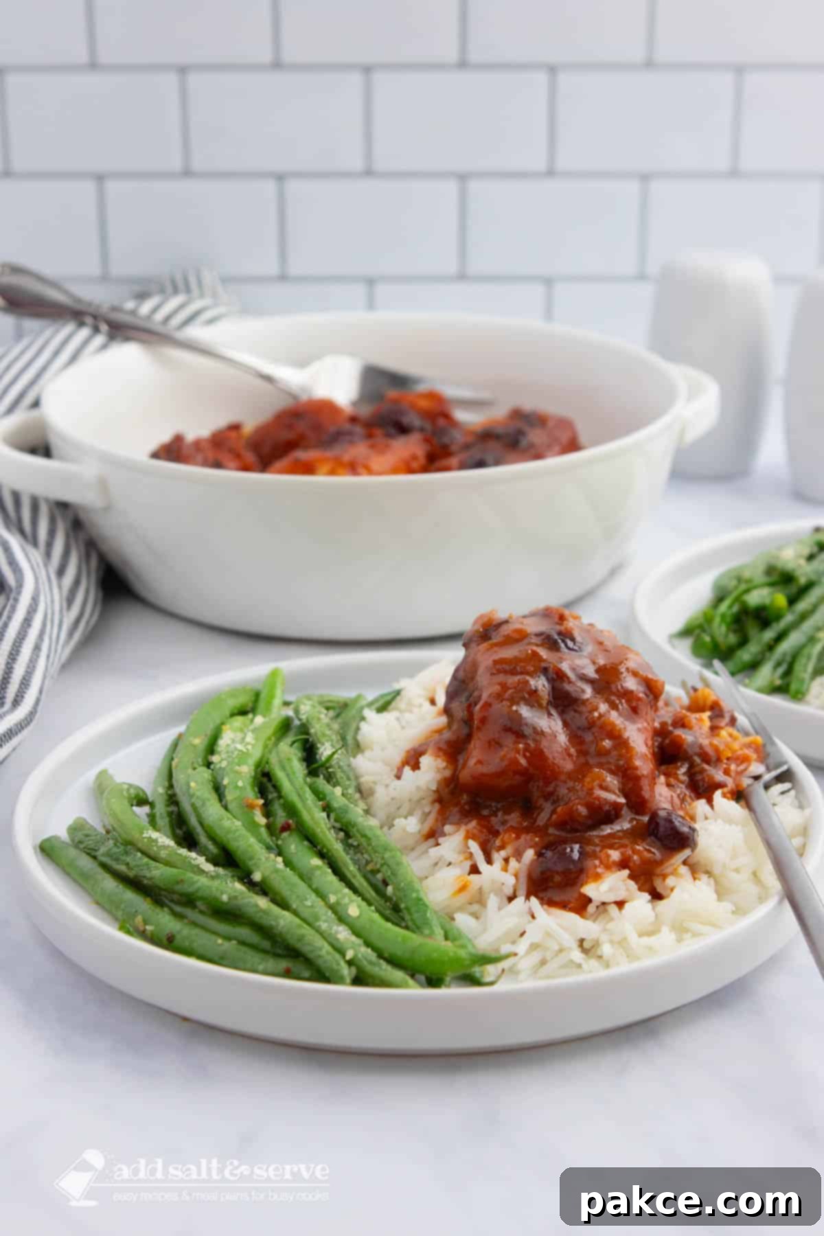 Slow Cooker Zesty Cranberry French Chicken 6 A white plate with seasoned green beans and chicken in a reddish-orange sauce with cranberries served over white rice with a serving dish of the chicken in the background.