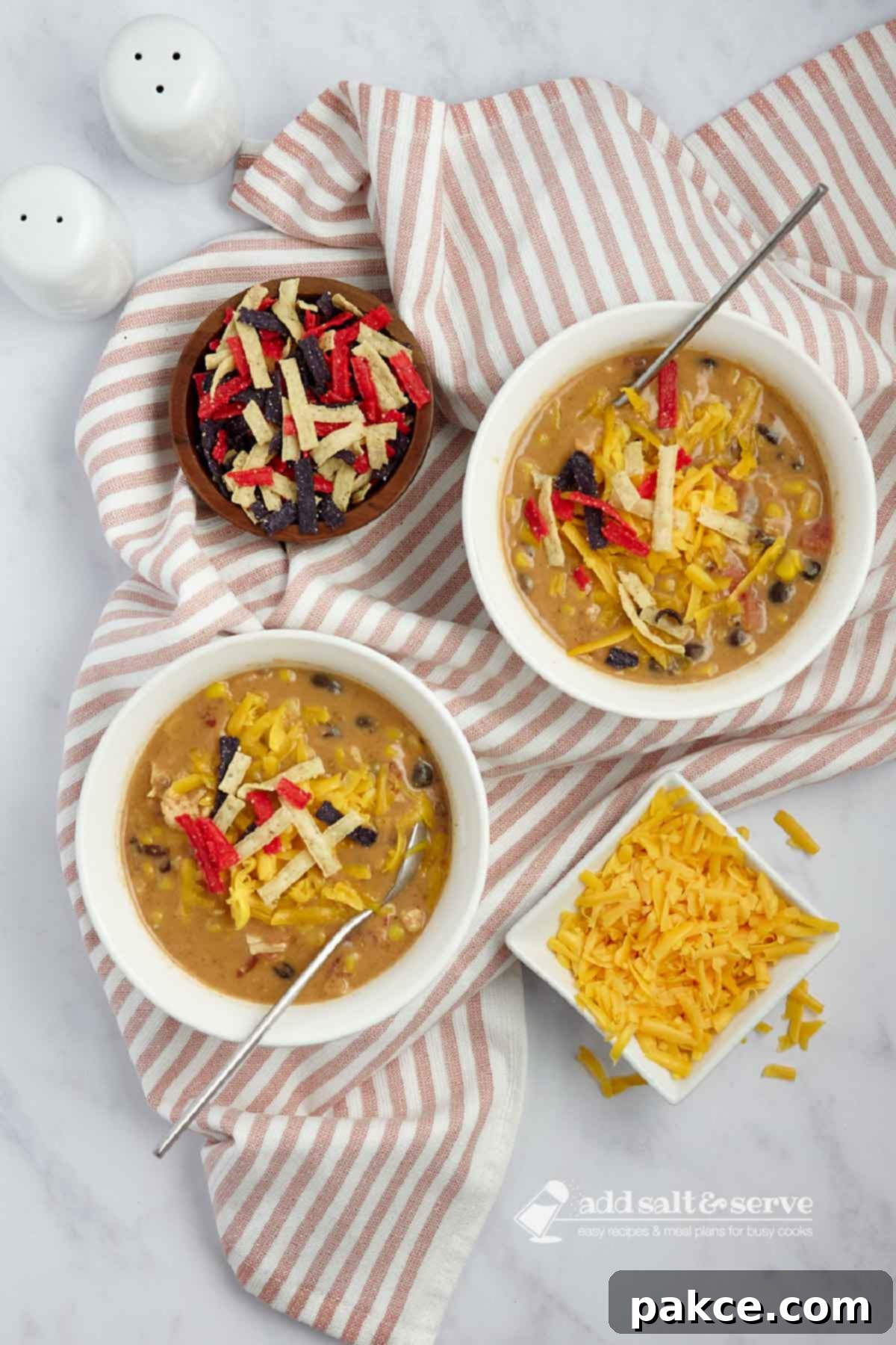 15 Minute Creamy Chicken Enchilada Soup 6 Two enticing bowls of creamy chicken enchilada soup are perfectly garnished with shredded cheddar cheese, vibrant cilantro, and crunchy tortilla strips. They are positioned beside small, elegant bowls holding additional shredded cheddar cheese and tortilla strips, inviting guests to add more to their liking.