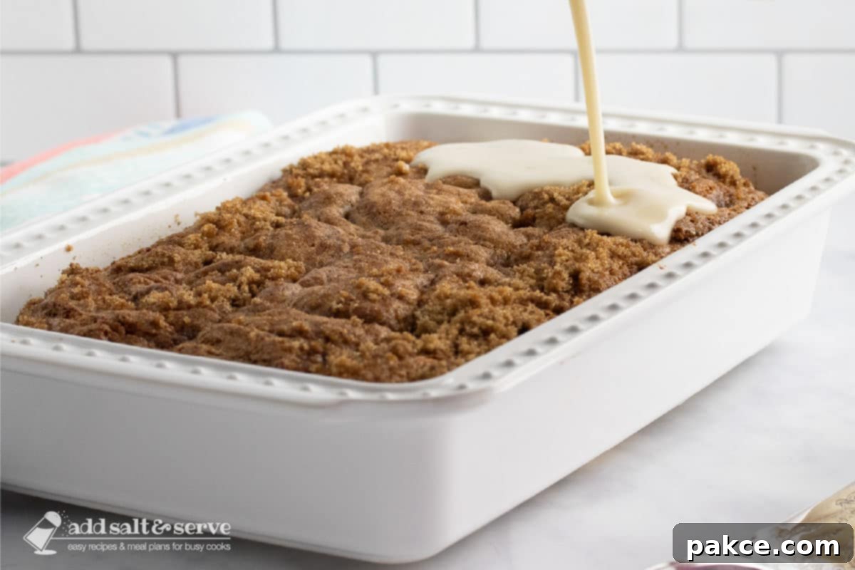 Honey-Glazed Cinnamon Swirl Cake 6 Baking pan with Honey Bun Cake with streusel topping with vanilla glaze being poured over the top.