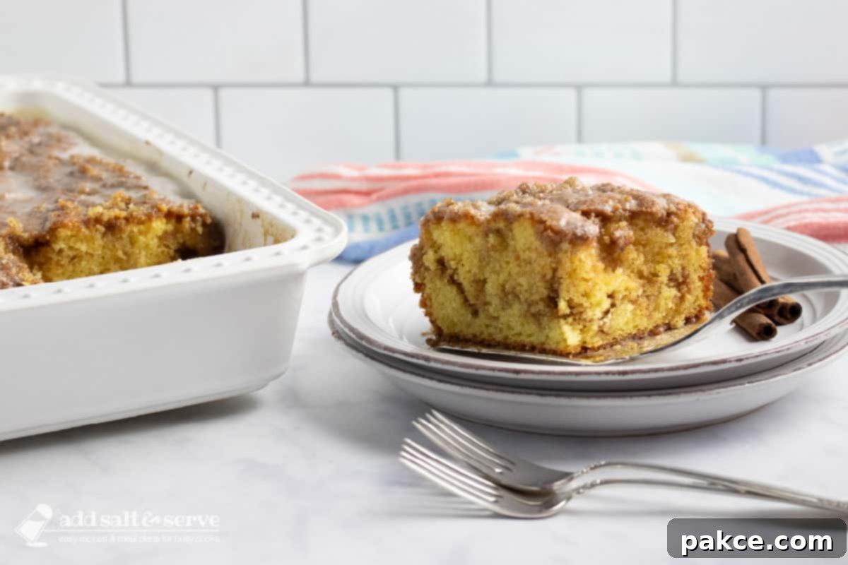 Honey-Glazed Cinnamon Swirl Cake 7 Slice of Honey Bun Cake being placed on a plate showing the cinnamon swirl in the middle and vanilla glaze on top on a counter beside the pan of cake and two forks.