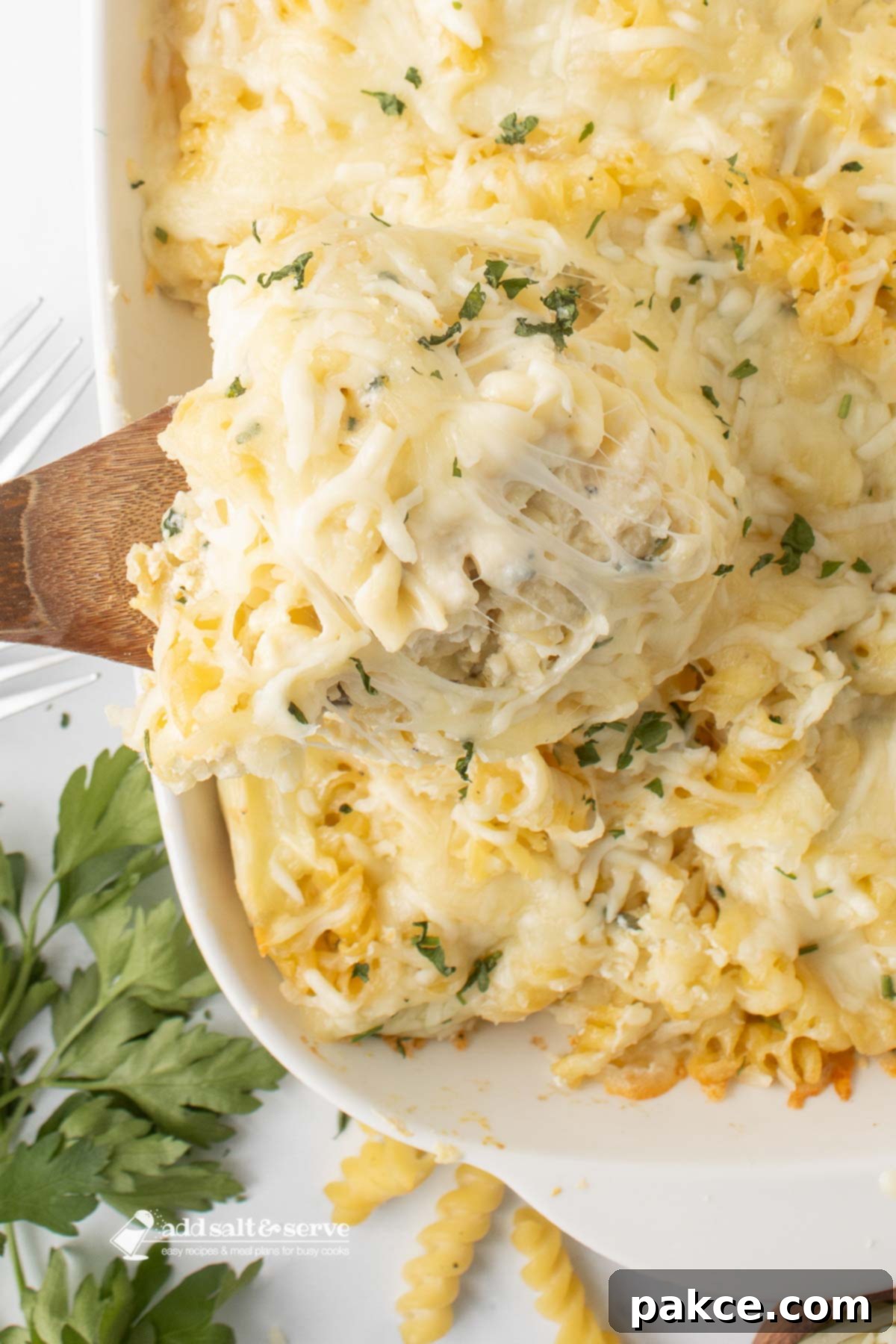 Creamy Alfredo Rotini Pasta Bake in a baking dish, with a serving scooped out onto a wooden spoon, showcasing its cheesy layers and spirals of pasta.