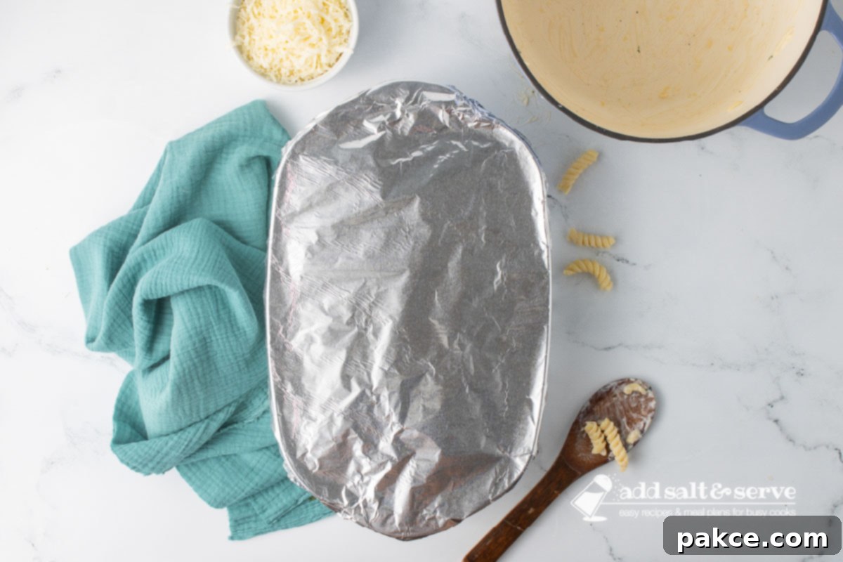 Creamy Alfredo Rotini Bake 10 An oblong baking dish covered tightly with aluminum foil, sitting on a kitchen counter next to an empty Dutch oven and a wooden spoon with leftover saucy rotini.