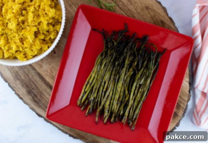 grilled asparagus on a red serving plate placed on a wood serving tray with a bowl of yellow rice and a tea towel to the side