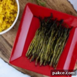 grilled asparagus on a red serving plate placed on a wood serving tray with a bowl of yellow rice and a tea towel to the side