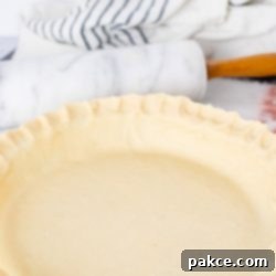 Simply Perfect Freezer Friendly Pie Crust 6 An unbaked pie crust beautifully crimped in a pie pan, with a marble rolling pin and a white linen towel in the soft background, highlighting the homemade quality.