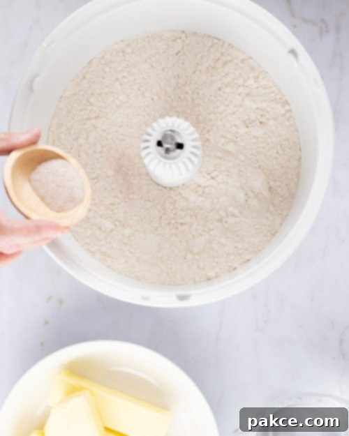 Overhead shot of flour in a mixer with salt being added from a small wooden bowl and containers of butter and water to the side, illustrating the initial mixing step for pie crust.