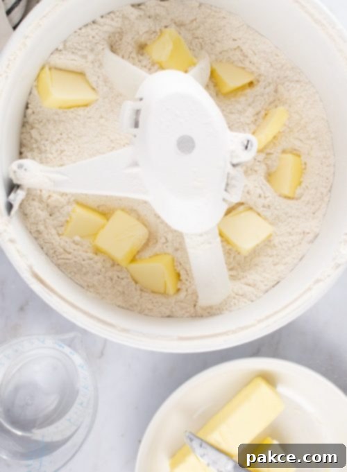 Overhead shot of flour in a mixer with chopped cold butter added throughout, a bowl with two sticks of butter to the side, and a measuring cup with 1-½ cups water to the side, showing the butter cutting process for flaky pie crust.