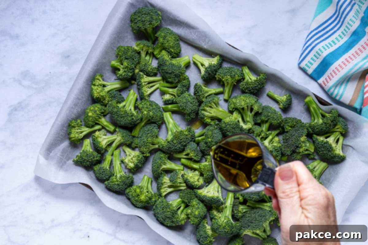 Garlic Roasted Broccoli 7 A person drizzles olive oil over broccoli florets on a parchment-lined baking sheet using a small measuring cup, preparing for seasoning.