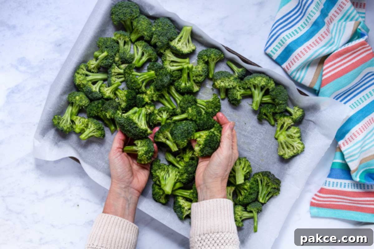 Garlic Roasted Broccoli 8 Hands are gently tossing broccoli florets with olive oil, garlic, salt, and pepper on a parchment-lined baking sheet to ensure even coating.