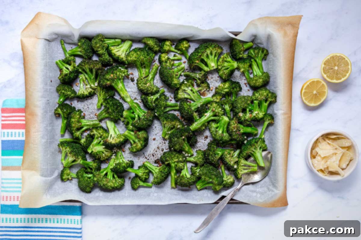 Garlic Roasted Broccoli 9 Freshly roasted broccoli florets on a parchment-lined sheet pan, golden and slightly charred, ready to be served. A small bowl of shaved Parmesan and lemon halves are positioned nearby for garnish.