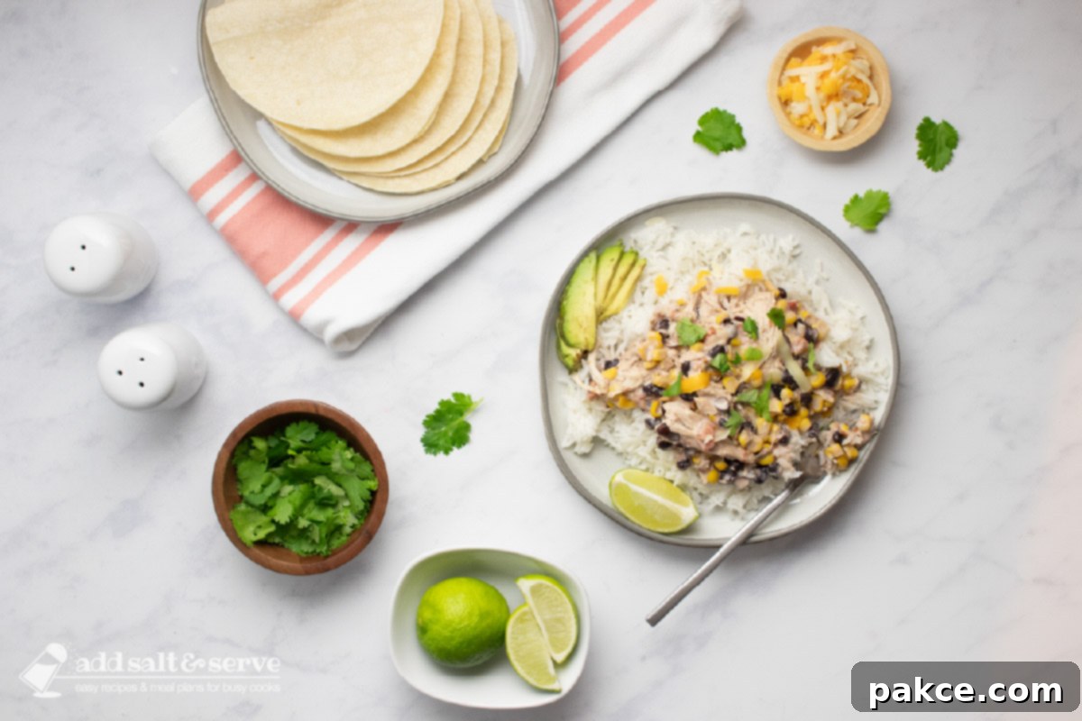 A plate of shredded chicken in a creamy sauce with black beans, corn, tomatoes, and green chilies served over rice with garnishes of sliced avocado, shredded cheese, fresh cilantro, and lime wedges and a plate of corn tortillas.