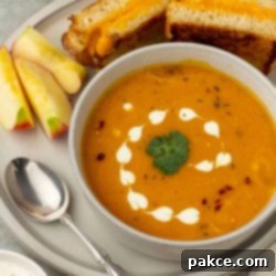 Golden Ginger Sweet Potato Carrot Soup 7 A bowl of vibrant, creamy carrot sweet potato ginger soup beautifully garnished with a swirl of sour cream and fresh cilantro, served with a grilled cheese sandwich and apple slices.