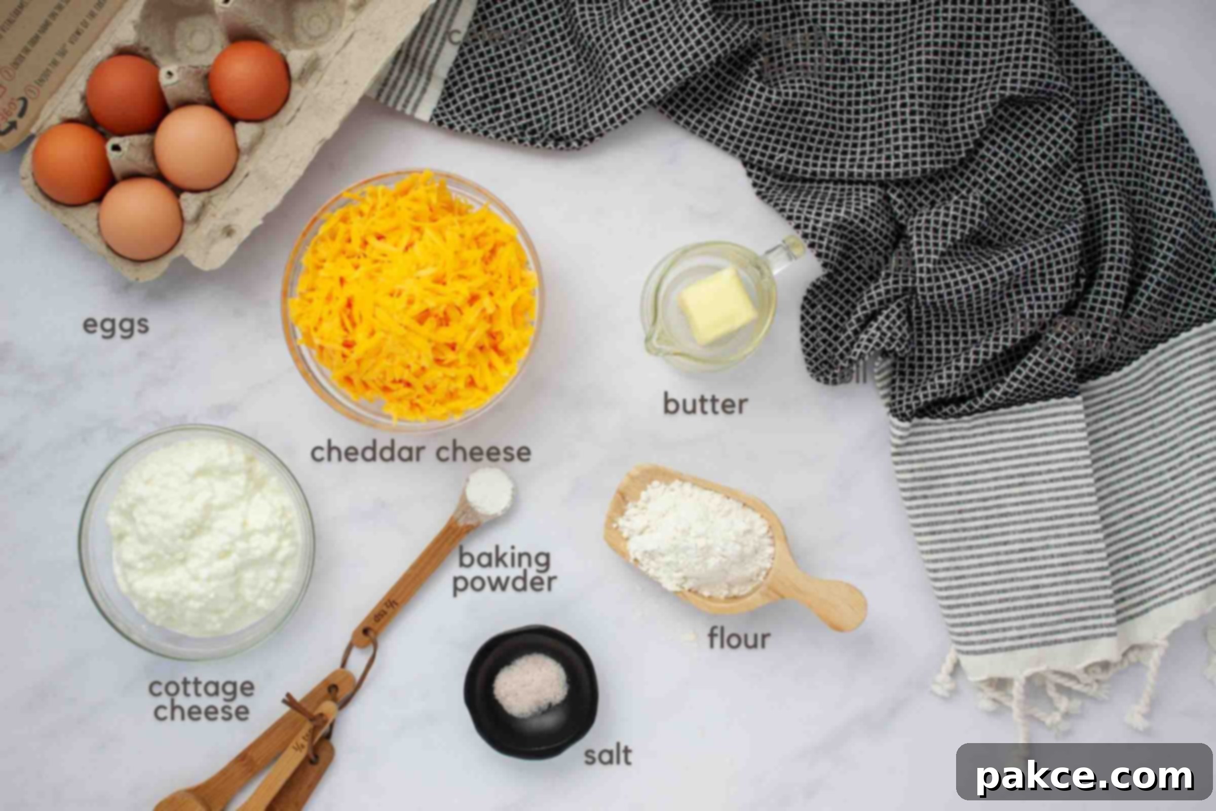 Fluffy Cottage Cheese Quiche 3 A selection of fresh ingredients neatly arranged on a kitchen counter, including eggs, shredded cheddar cheese, cottage cheese, baking powder, salt, flour, and a stick of butter, all ready for making a crustless quiche.