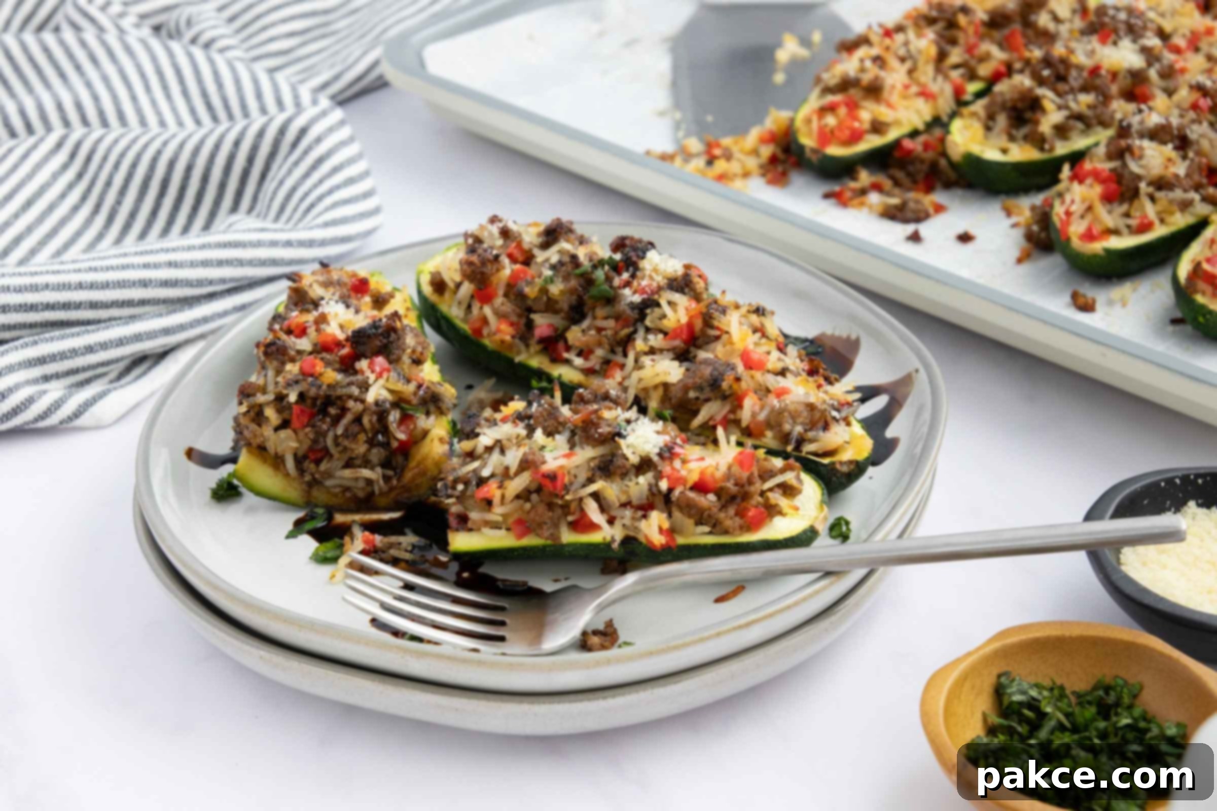 Baked zucchini stuffed with a rice, ground beef, and vegetable mixture drizzled with balsamic glaze on a plate next to a baking sheet lined with parchment paper with more zucchini.