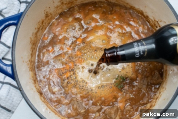 Beef and Guinness Stew Guinness beer being poured from a bottle into a pot of beef stew with onions and carrots, creating a rich base.