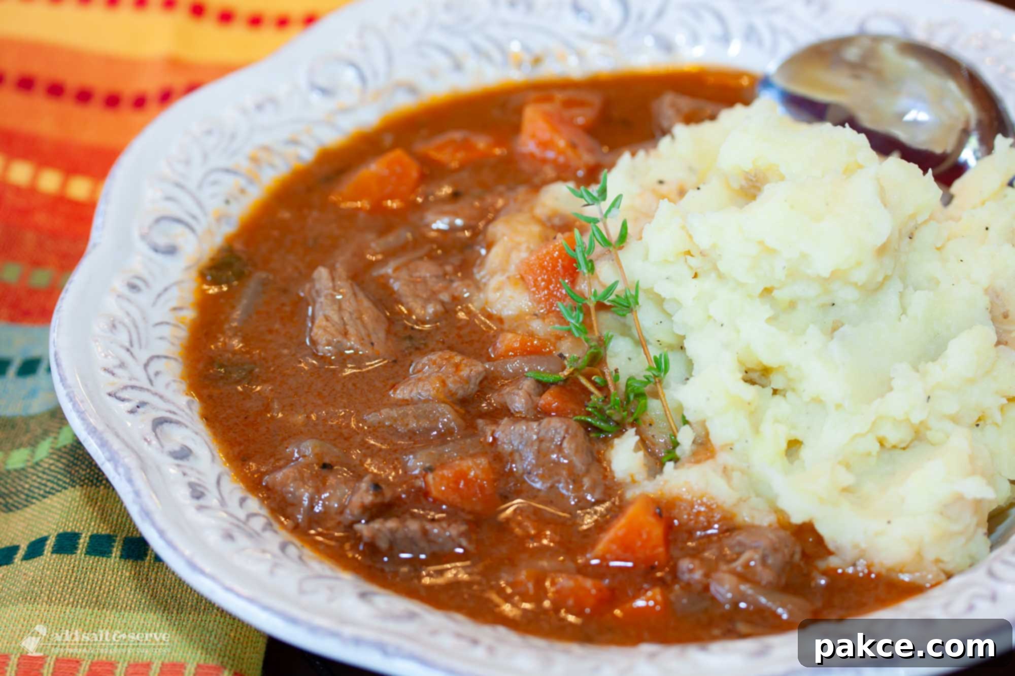 Hearty and Hassle-Free Beef and Guinness Stew 2 A beautifully presented white bowl of beef stew with tender beef chunks and carrots, served over a bed of fluffy mashed potatoes and garnished with a sprig of fresh thyme.
