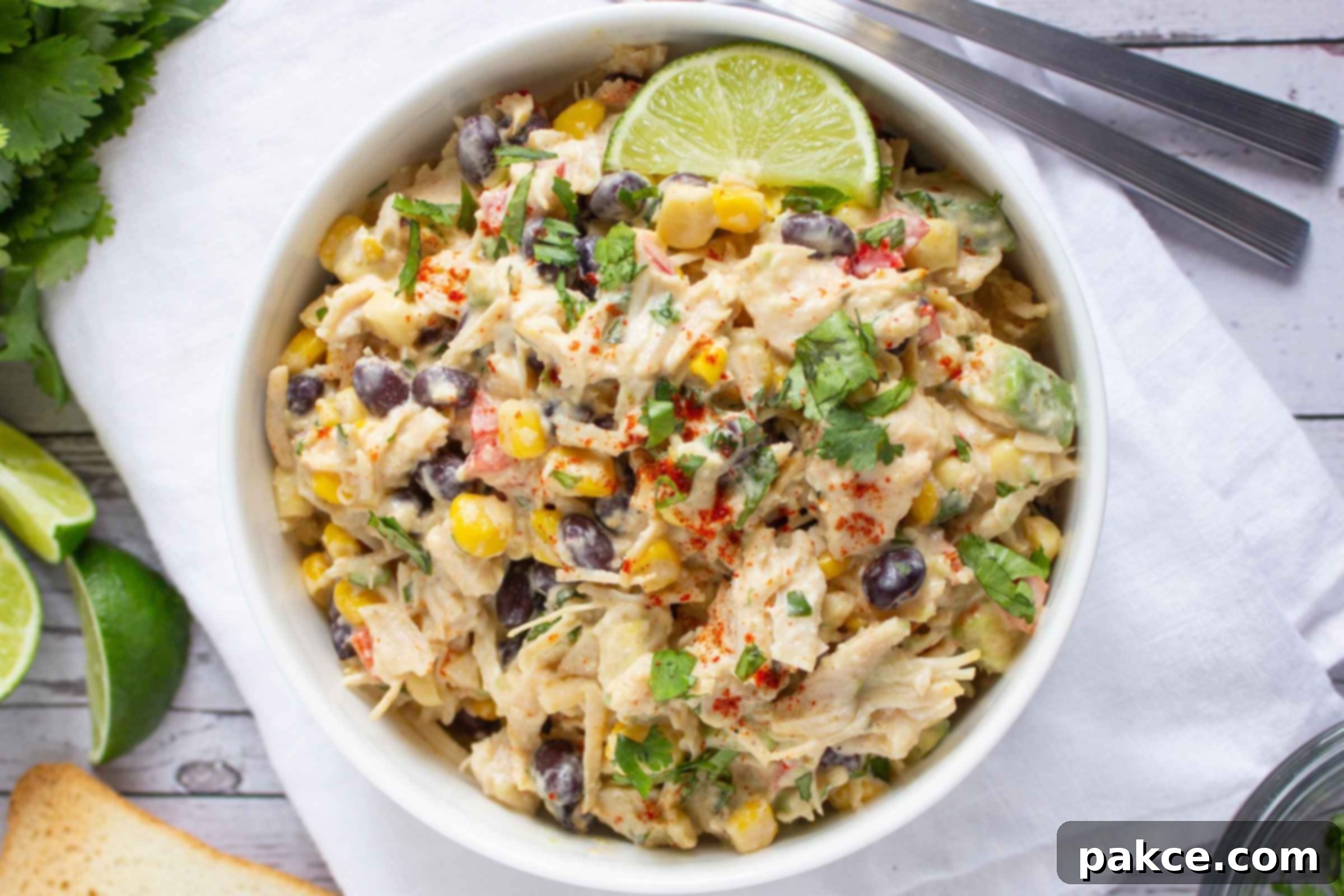 Bowl of chicken salad with corn, black beans, avocado, and red bell pepper in a creamy dressing garnished with a lime wedge and sprinkled with paprika.