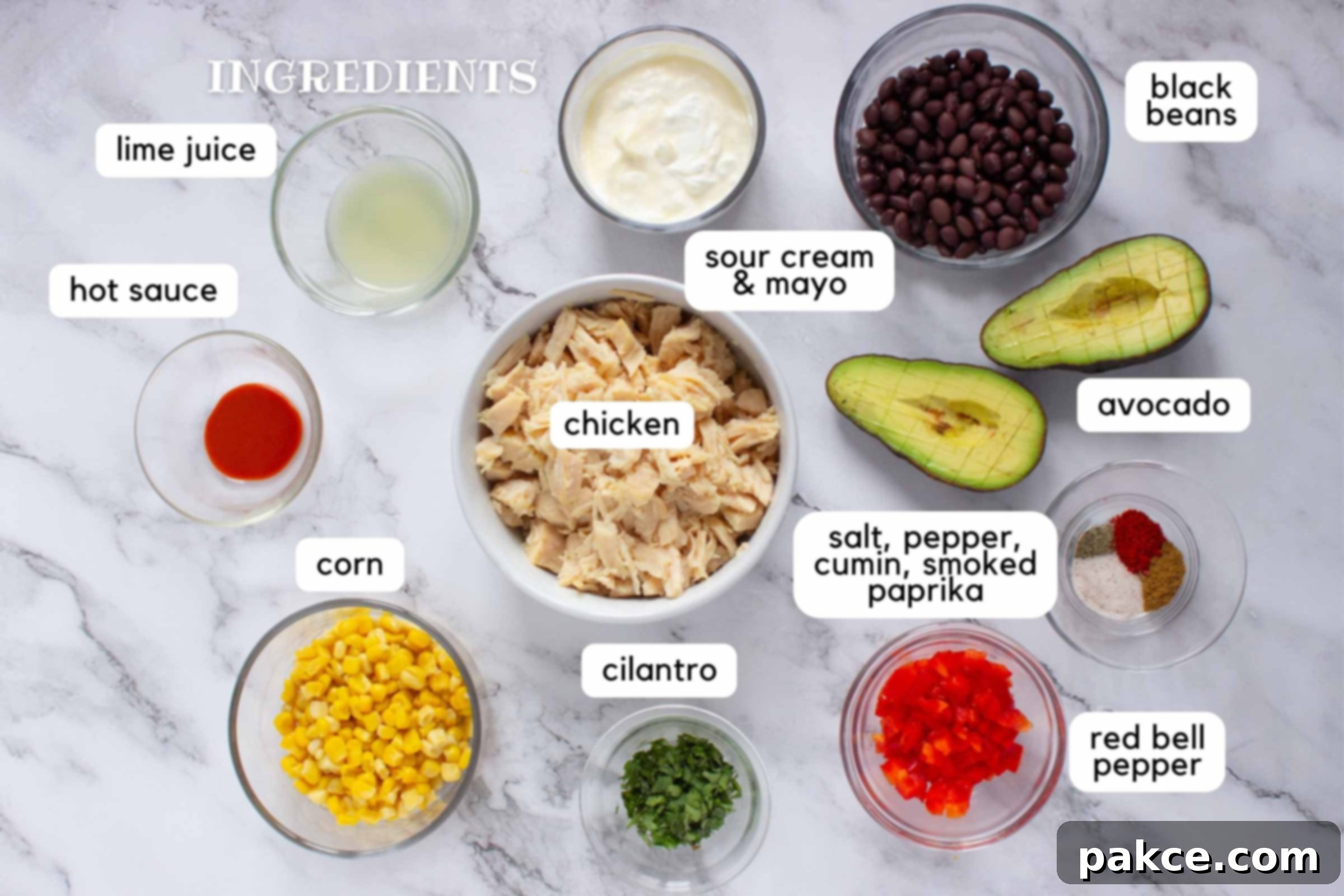 Fiesta Lime Chicken Salad 3 Labeled ingredients on a counter: Chicken, corn, cilantro, salt, pepper, cumin, smoked paprika, red bell pepper, avocado, black beans, sour cream & mayo, lime juice, and hot sauce.