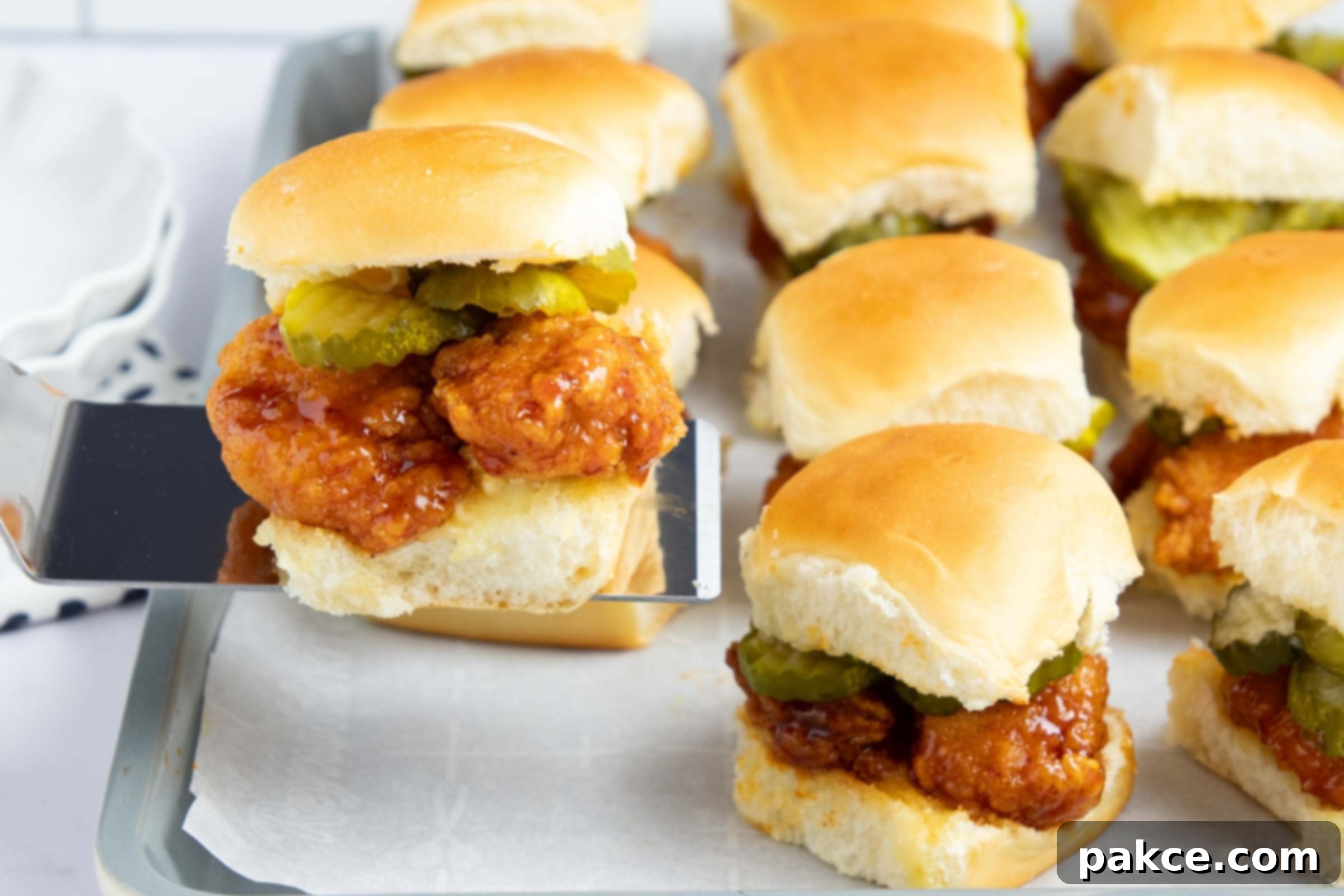 Hot honey chicken sliders with pickles on a baking sheet with one lifted on a spatula.