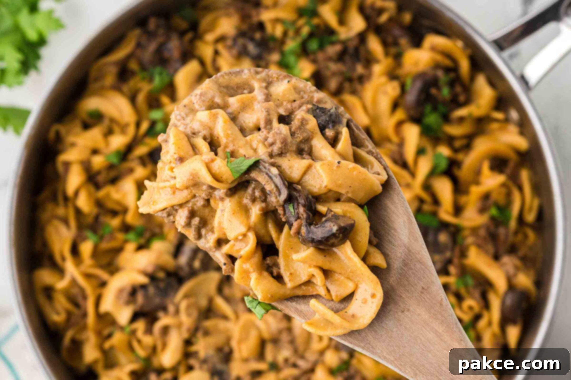 A pan with a finished recipe showing egg noodles, sliced mushrooms, and ground beef in a sauce garnished with chopped parsley with a serving lifted toward the camera on a wooden spoon.