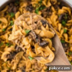 Quick and Creamy One-Pot Ground Beef Stroganoff 11 A pan with a finished recipe showing egg noodles, sliced mushrooms, and ground beef in a sauce garnished with chopped parsley with a serving lifted toward the camera on a wooden spoon.