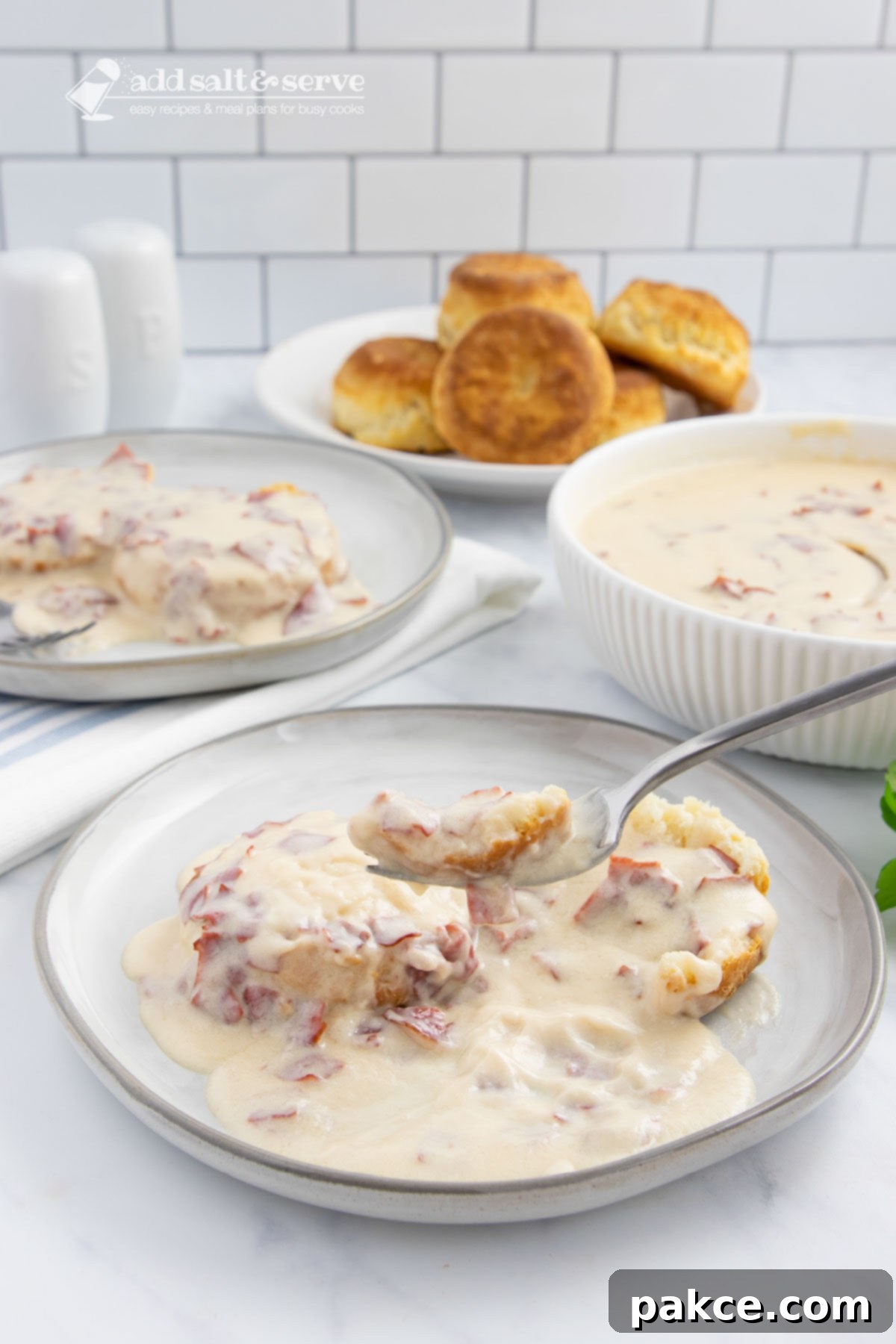 Hearty Chipped Beef Gravy 7 A close-up of a plate of creamy chipped beef gravy over two biscuit halves, with a fork lifting a spoonful of gravy, set against a backdrop of more gravy and biscuits.
