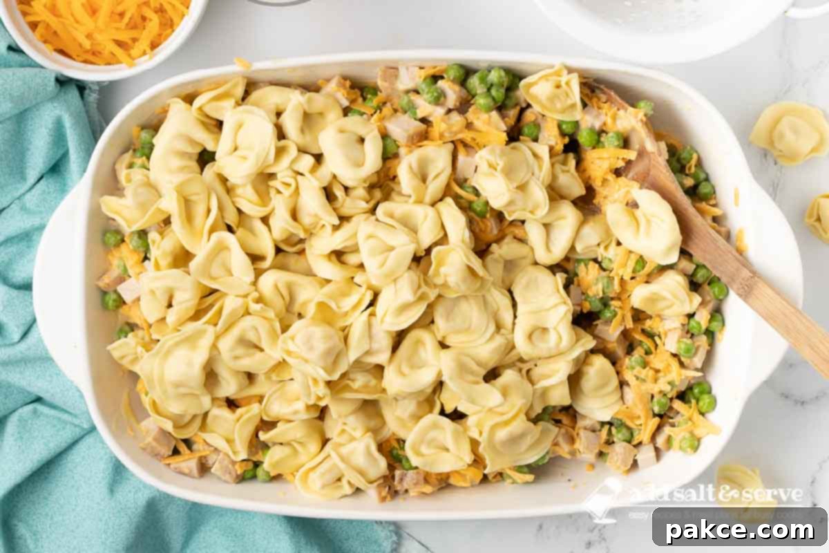 Creamy Turkey Tortellini Bake 6 Cooked and drained cheese tortellini being added into a large casserole dish, which already contains the creamy mixture of leftover chopped turkey, peas, and cheese sauce. A spatula is poised for gentle mixing.
